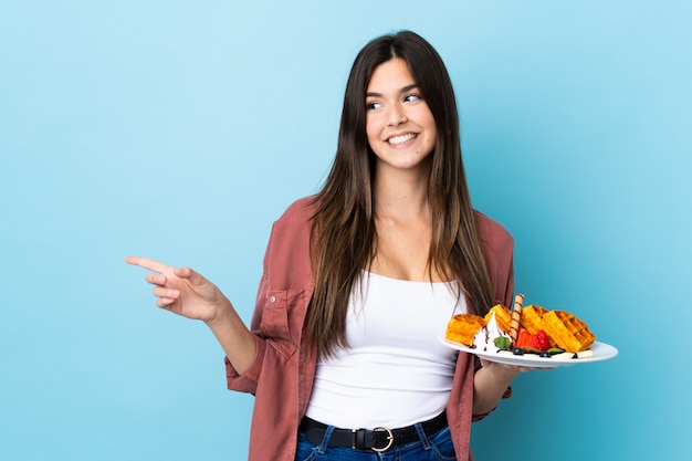 Teenager Brazilian girl holding waffles over isolated blue wall pointing to the side to present a product