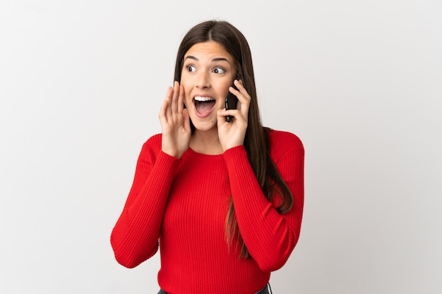 Teenager brazilian girl using mobile phone over isolated white background shouting with mouth wide open to the side