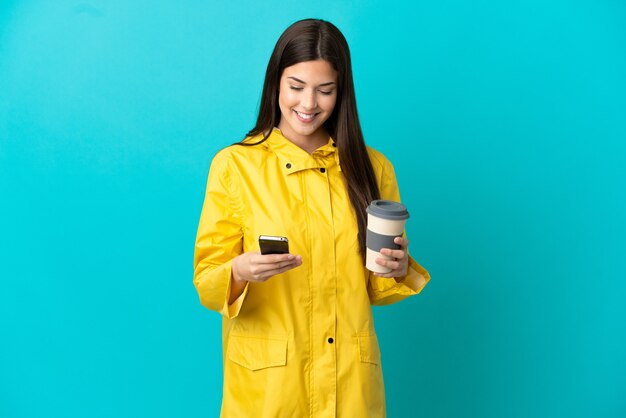 Teenager Brazilian girl wearing a rainproof coat over isolated blue background holding coffee to take away and a mobile