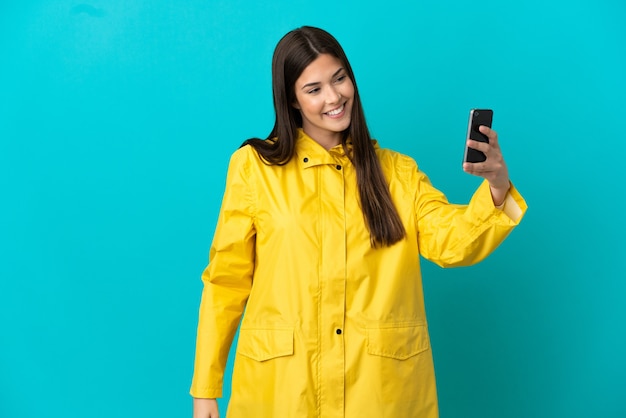 Teenager Brazilian girl wearing a rainproof coat over isolated blue background making a selfie