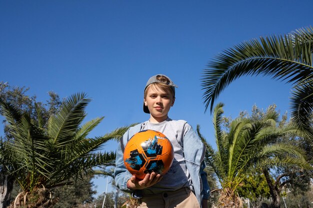 Teenager in cap and casual clothes holds orange basketball\
stands against background of blue sky and palm trees looks at\
camera sport active leisure time concept summer games copy\
space