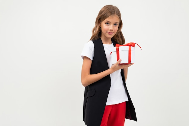 Teenager caucasian girl in black jacket, white shirt and red pants holds a white box with gift and rejoices isolated on white