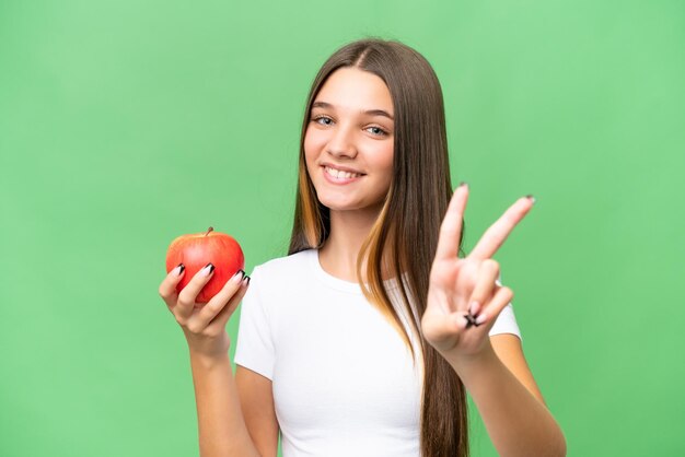 Teenager caucasian girl holding an apple over isolated background smiling and showing victory sign