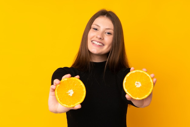 Teenager caucasian girl holding an orange over isolated yellow wall