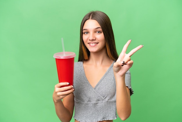 Teenager caucasian girl holding a soda over isolated background smiling and showing victory sign