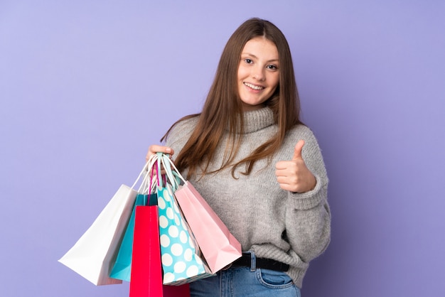 Teenager caucasian girl isolated on purple wall holding shopping bags and with thumb up