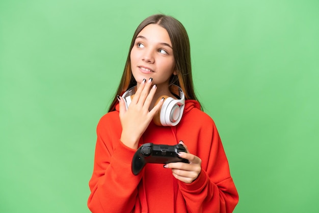 Teenager caucasian girl playing with a video game controller over isolated background looking up while smiling