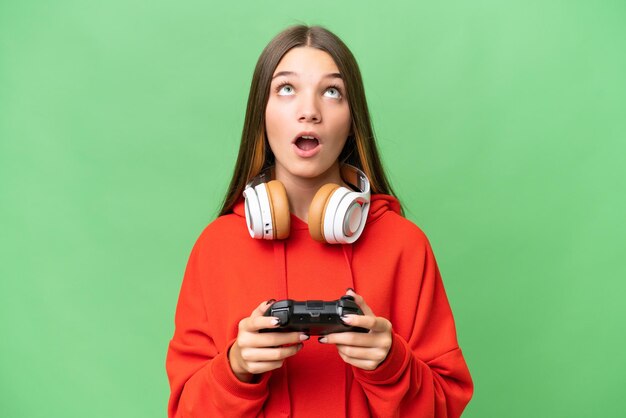 Teenager caucasian girl playing with a video game controller over isolated background looking up and with surprised expression