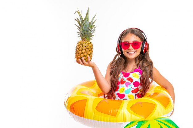 Teenager caucasian girl in sunglasses with pineapple and rubber mattresses on white wall