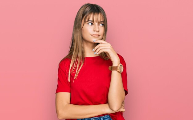 Foto adolescente ragazza caucasica che indossa una maglietta rossa casual con la mano sul mento che pensa alla domanda espressione pensierosa sorridente con il concetto di dubbio viso premuroso