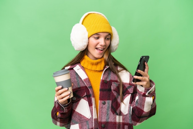 외진 배경 위에 겨울 머프를 입은 10대 백인 소녀가 커피를 들고 휴대전화를 들고 있습니다.