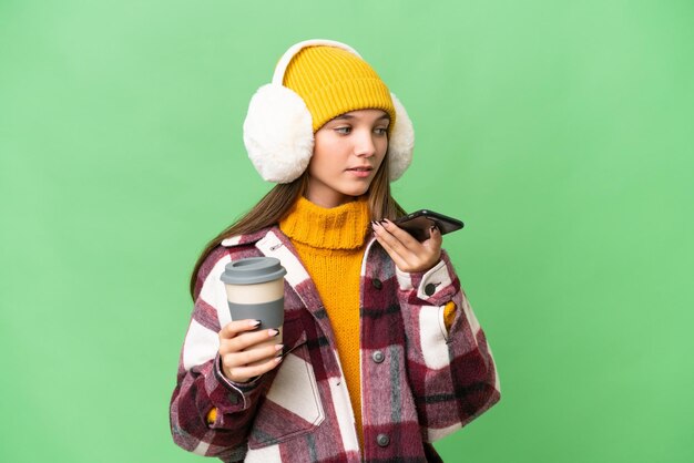 Teenager caucasian girl wearing winter muffs over isolated background holding coffee to take away and a mobile