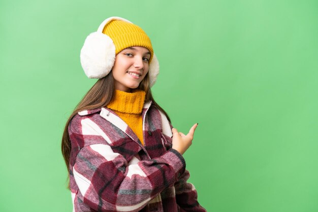 Teenager caucasian girl wearing winter muffs over isolated background pointing back