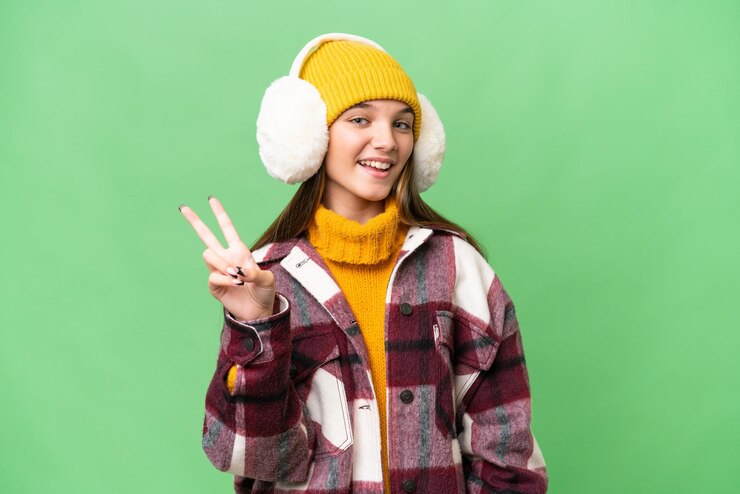 Photo teenager caucasian girl wearing winter muffs over isolated background smiling and showing victory sign