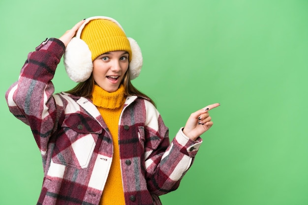 Кавказская девушка-подросток в зимних муфтах на изолированном фоне удивлена и показывает пальцем в сторону