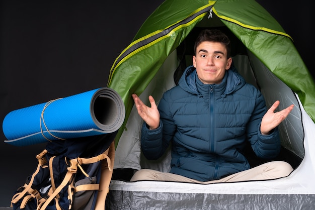 Uomo caucasico dell'adolescente dentro una tenda verde di campeggio sulla parete nera che ha dubbi mentre sollevando le mani