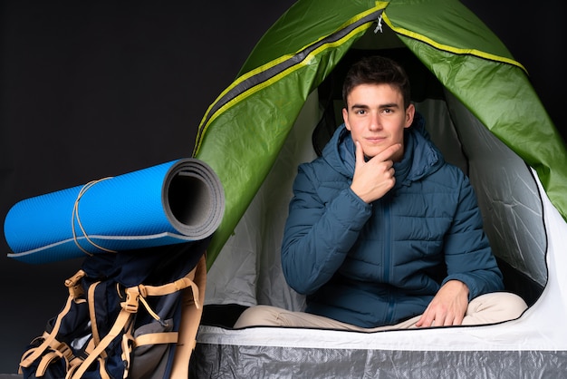 Teenager caucasian man inside a camping green tent on black wall thinking