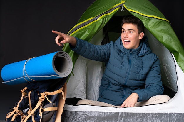 Teenager caucasian man inside a camping green tent isolated on black pointing away