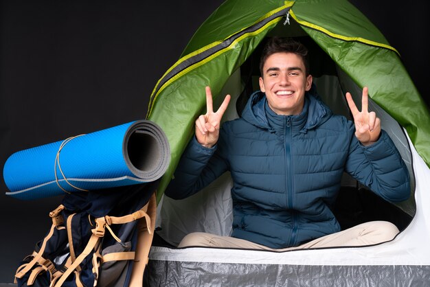 Teenager caucasian man inside a camping green tent isolated on black showing victory sign with both hands