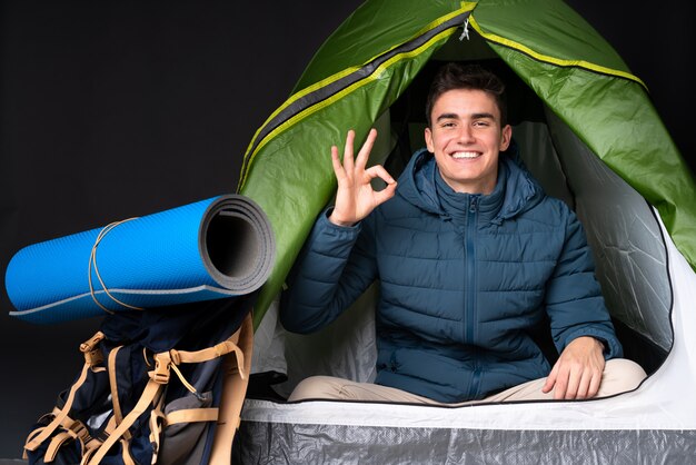 Foto uomo caucasico dell'adolescente dentro una tenda verde di campeggio isolata sulla parete nera che mostra segno giusto con le dita