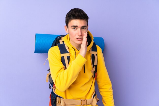 Teenager caucasian mountaineer man with a big backpack isolated on purple background whispering something