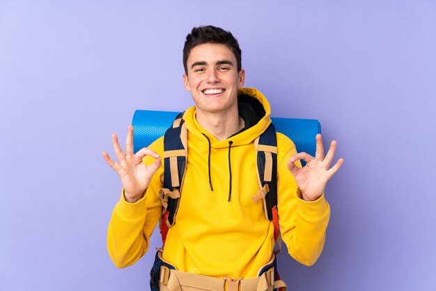 Teenager caucasian mountaineer man with a big backpack isolated on purple wall showing an ok sign with fingers