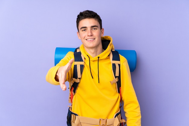 Teenager caucasian mountaineer man with a big backpack on purple wall handshaking after good deal