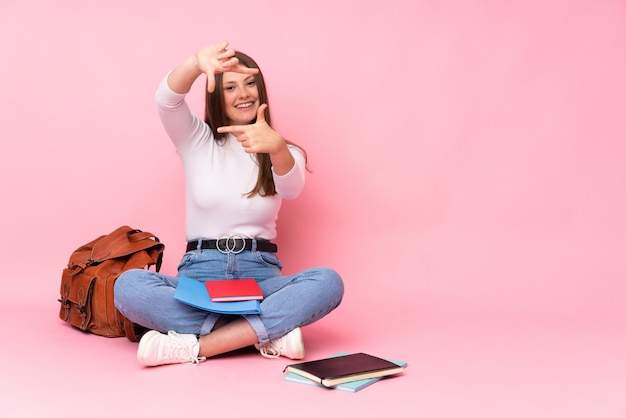 Teenager caucasian student girl sitting on the floor isolated on pink focusing face. Framing symbol