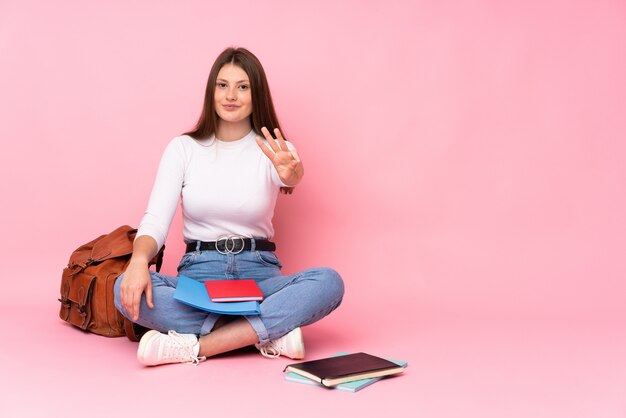 Ragazza caucasica dello studente dell'adolescente che si siede sul pavimento isolato sul rosa felice e che conta tre con le dita