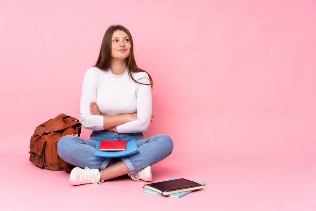 Teenager caucasian student girl sitting on the floor isolated on pink looking up while smiling