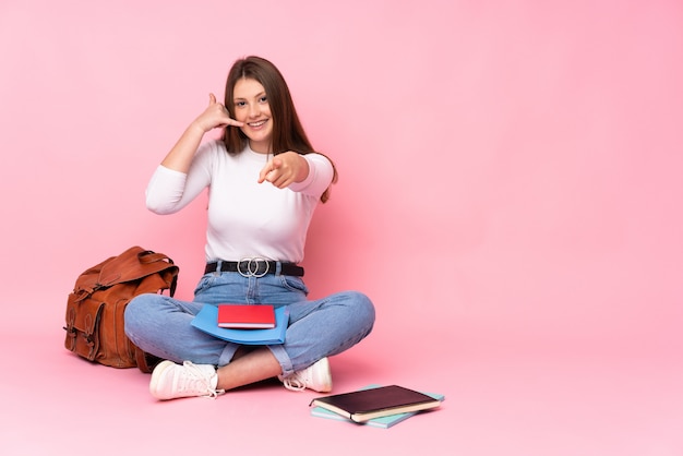 Teenager caucasian student girl sitting on the floor isolated on pink making phone gesture and pointing front