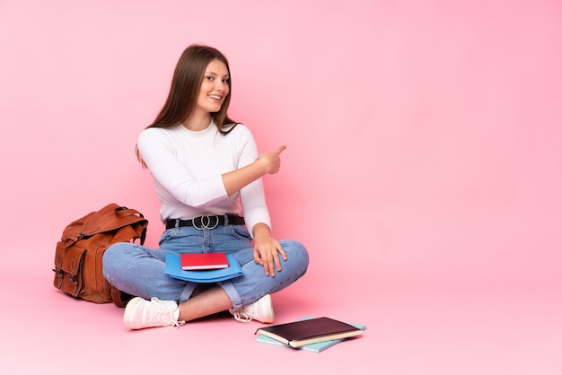 Teenager caucasian student girl sitting on the floor isolated on pink pointing back