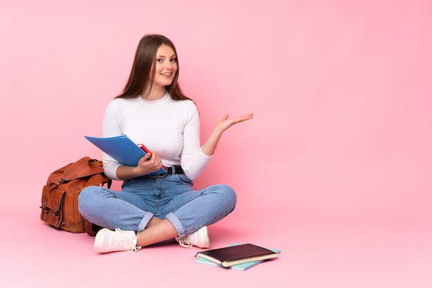 Ragazza caucasica dello studente dell'adolescente che si siede sul pavimento isolato sul rosa che presenta un'idea mentre guardando sorridendo verso