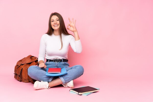 Teenager caucasian student girl sitting on the floor isolated on pink surprised and showing ok sign