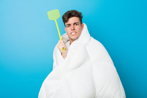 Teenager caucasian tired man covering himself with a blanket and holding a flyswatter