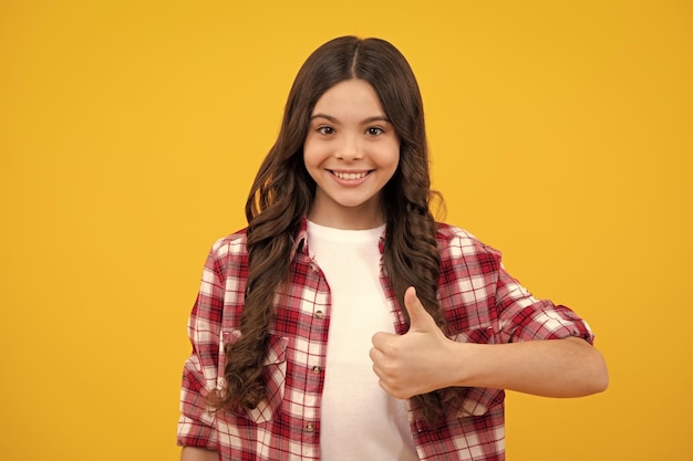 Teenager child girl 12 13 14 year old doing thumbs up gesture Cheerful teenager girl showing thumbs up sign isolated on yellow background Happy teenager positive and smiling emotions