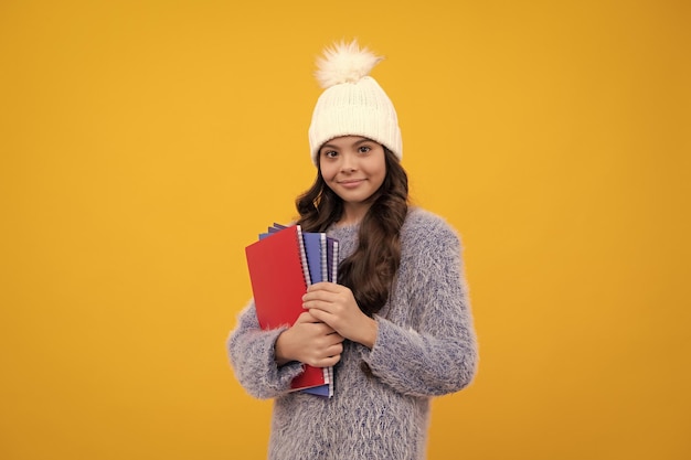 Teenager child in warm sweater and hat hold books and backpack isolated on yellow background happy schoolgirl positive and smiling emotions