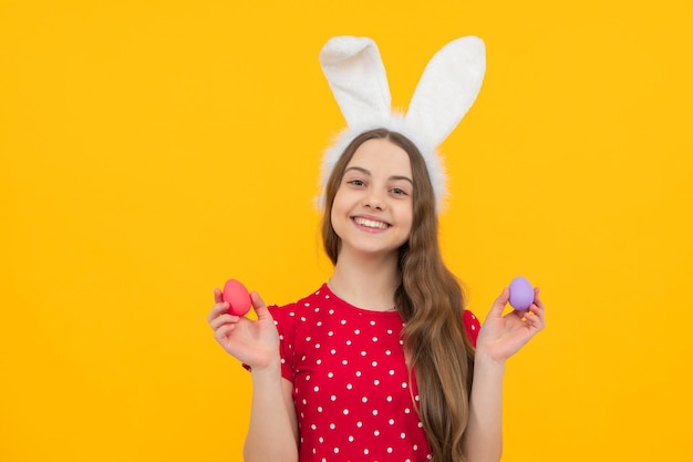 Ребенок-подросток в кроличьих ушах и держит красочные расписные пасхальные яйца на желтом фоне