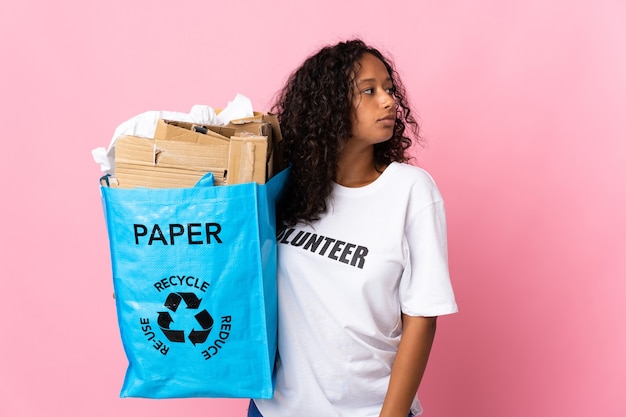 Teenager cuban girl holding a recycling bag full of paper to recycle isolated on pink background looking to the side