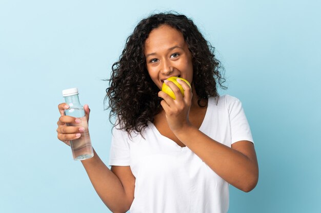 Teenager cuban girl isolated on blue wall with a bottle of water and eating an apple