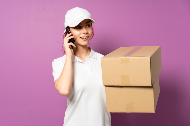 Teenager delivery woman isolated on purple wall keeping a conversation with the mobile phone with someone