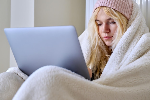 Teenager frozen in winter cold season warming up with blanket hat looking at laptop