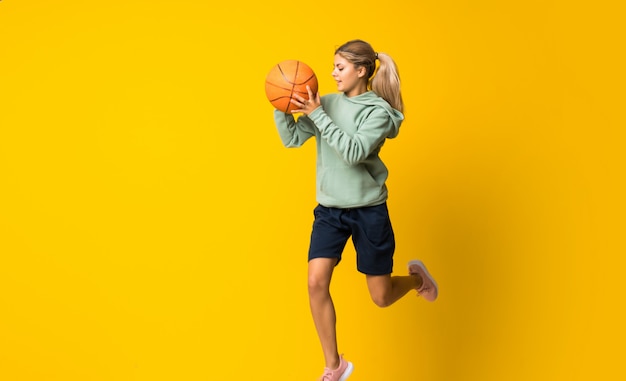 Teenager girl basketball ball jumping over isolated yellow wall