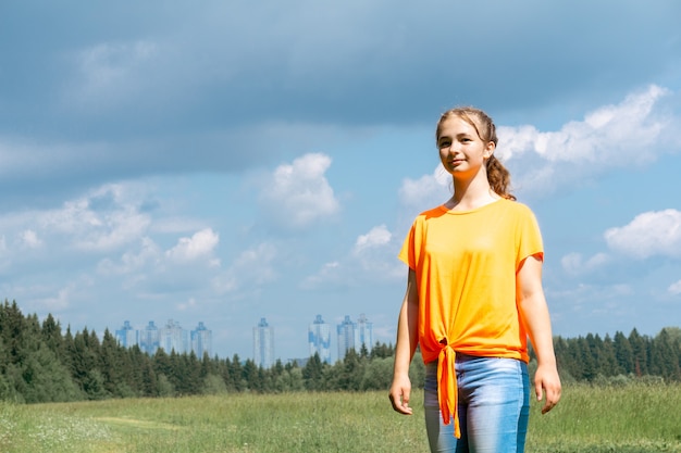 明るいオレンジ色のTシャツを着た10代の少女は、遠くの街を背景に自然の中を歩きます