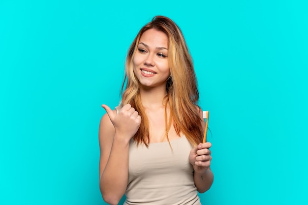 Teenager girl brushing teeth over isolated blue background pointing to the side to present a product