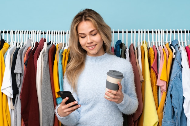 Teenager girl buying some clothes isolated on blue wall holding coffee to take away and a mobile