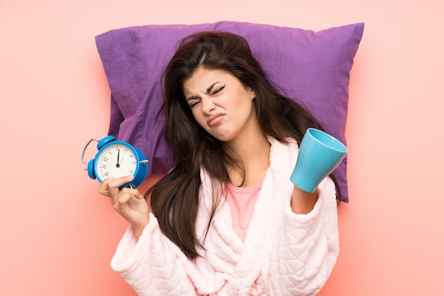 Teenager girl in dressing gown over pink backgrounnd and\
stressed holding vintage clock and holding a cup of coffee