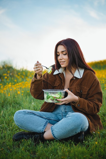 Photo teenager girl eating a salad while sitting on grass at a hill surrounded by yellow flowers