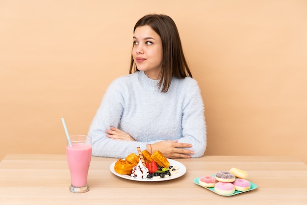 Teenager girl eating waffles on beige wall making doubts gesture looking side