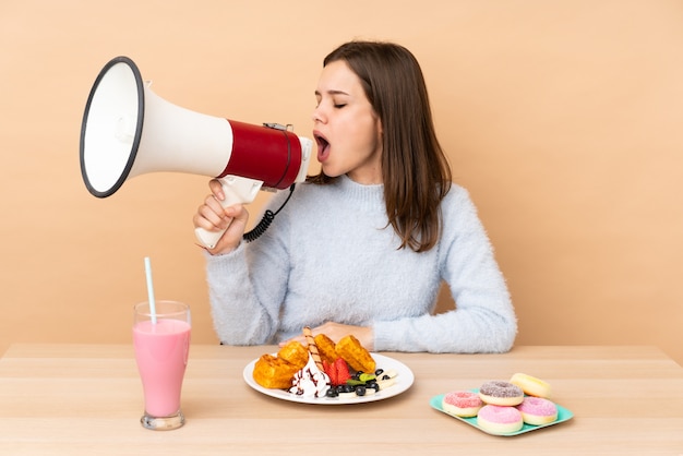 Teenager girl eating waffles isolated on beige wall shouting through a megaphone